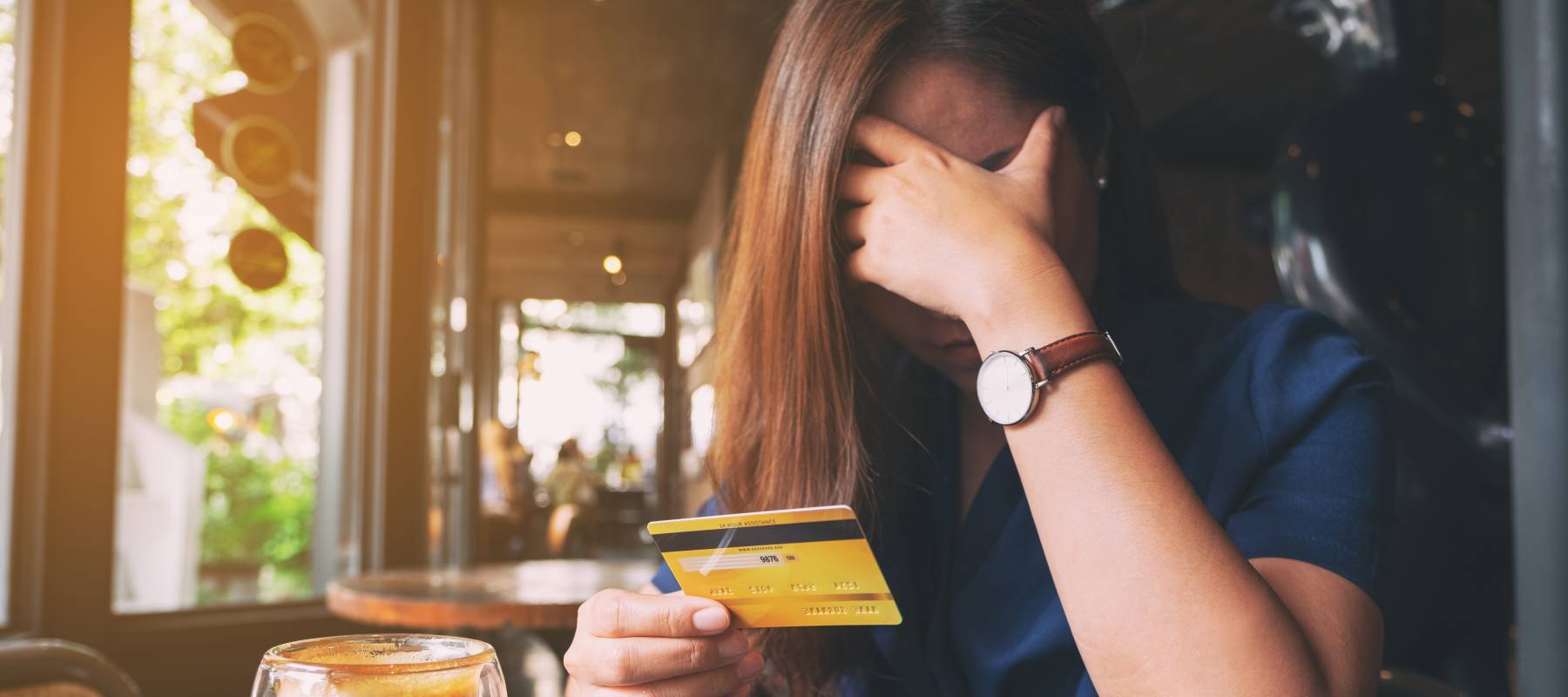 Close up of a woman in a coffee shop holding her credit card in one hand and the other covering her face, looking distressed.