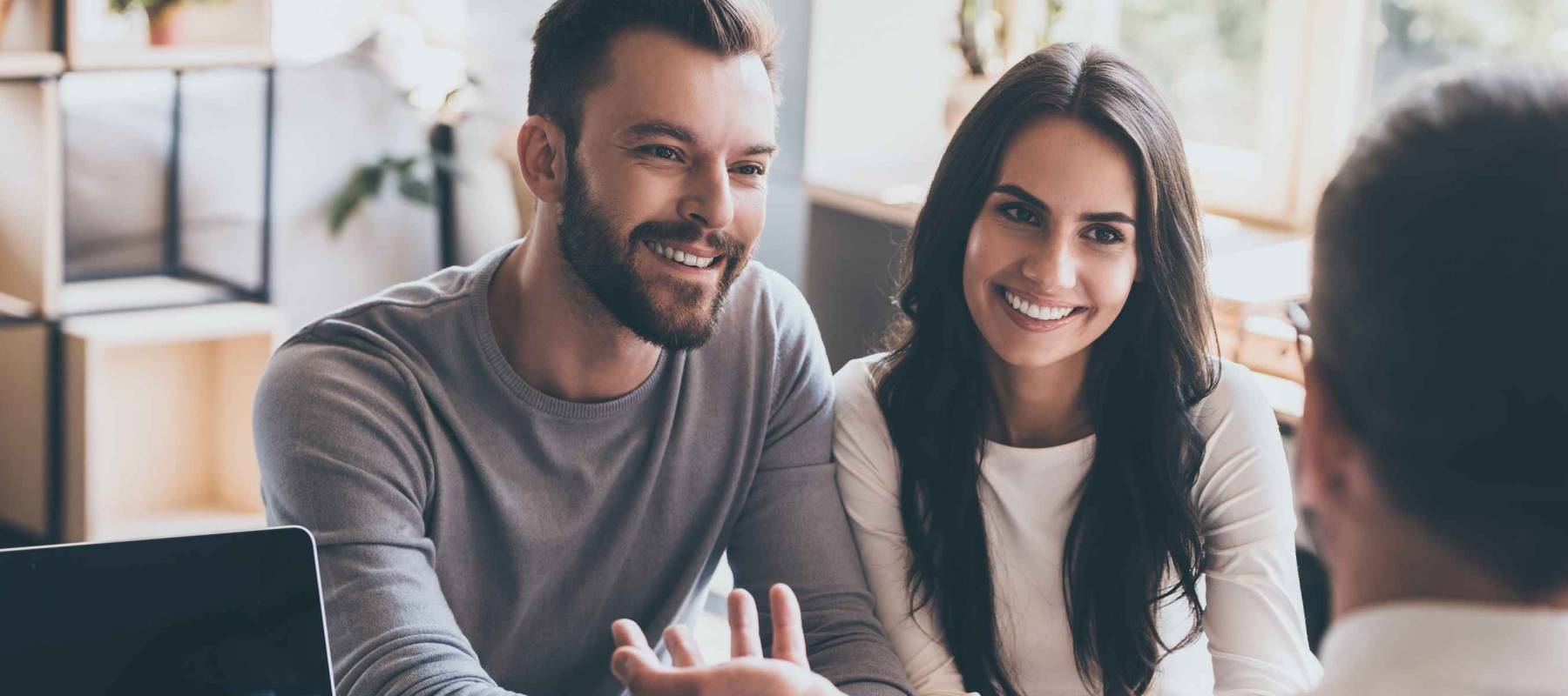 Good news. Happy young loving couple bonding to each other and looking at man sitting in front of them at the desk and gesturing