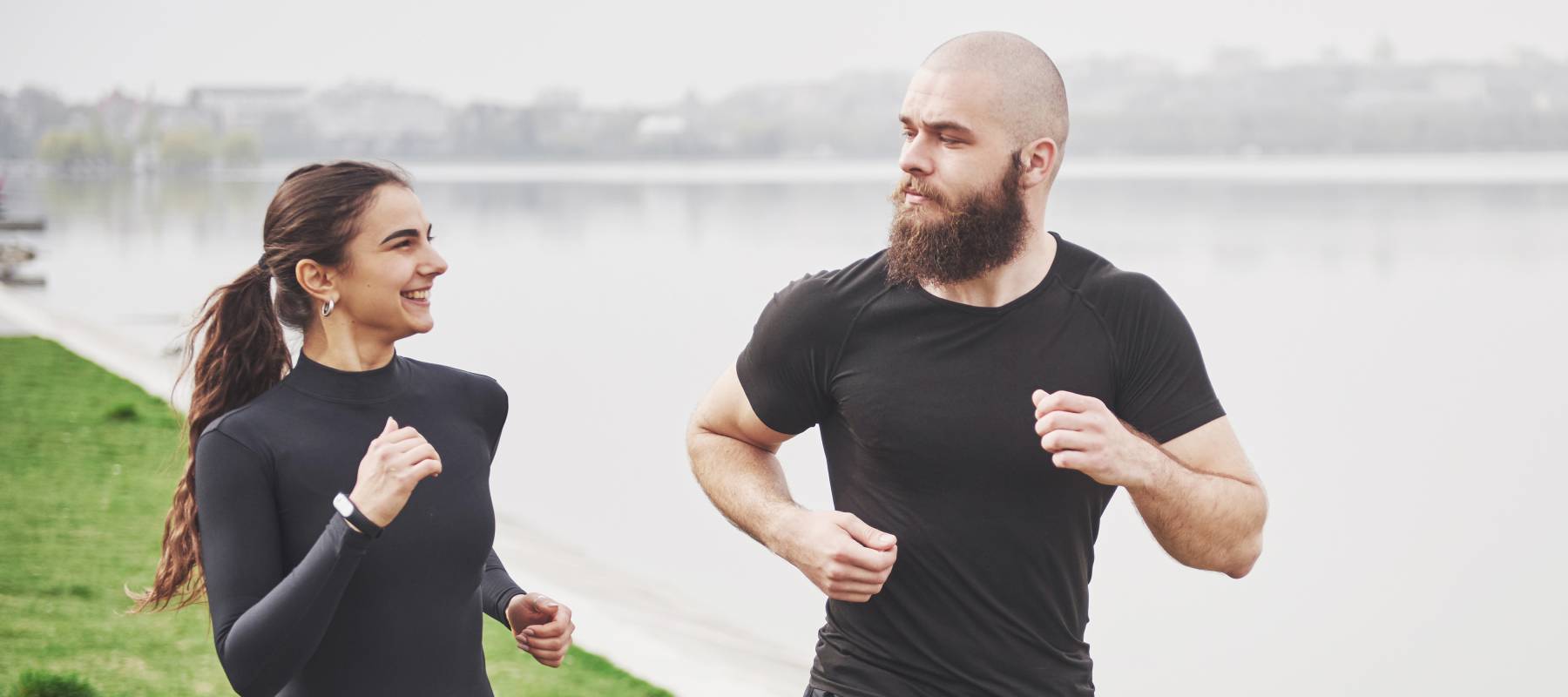 A couple in black running gear jog along a river.