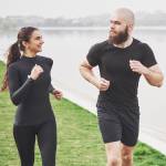 A couple in black running gear jog along a river.