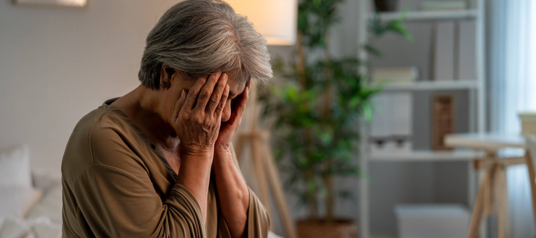 A sad older woman is sitting alone at home with her face in her hands.