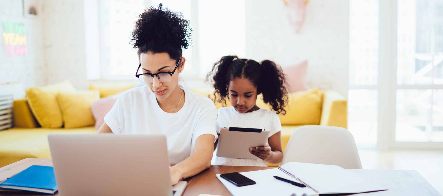 African American little girl standing in bright room and browsing tablet while mother in glasses working and typing on laptop
