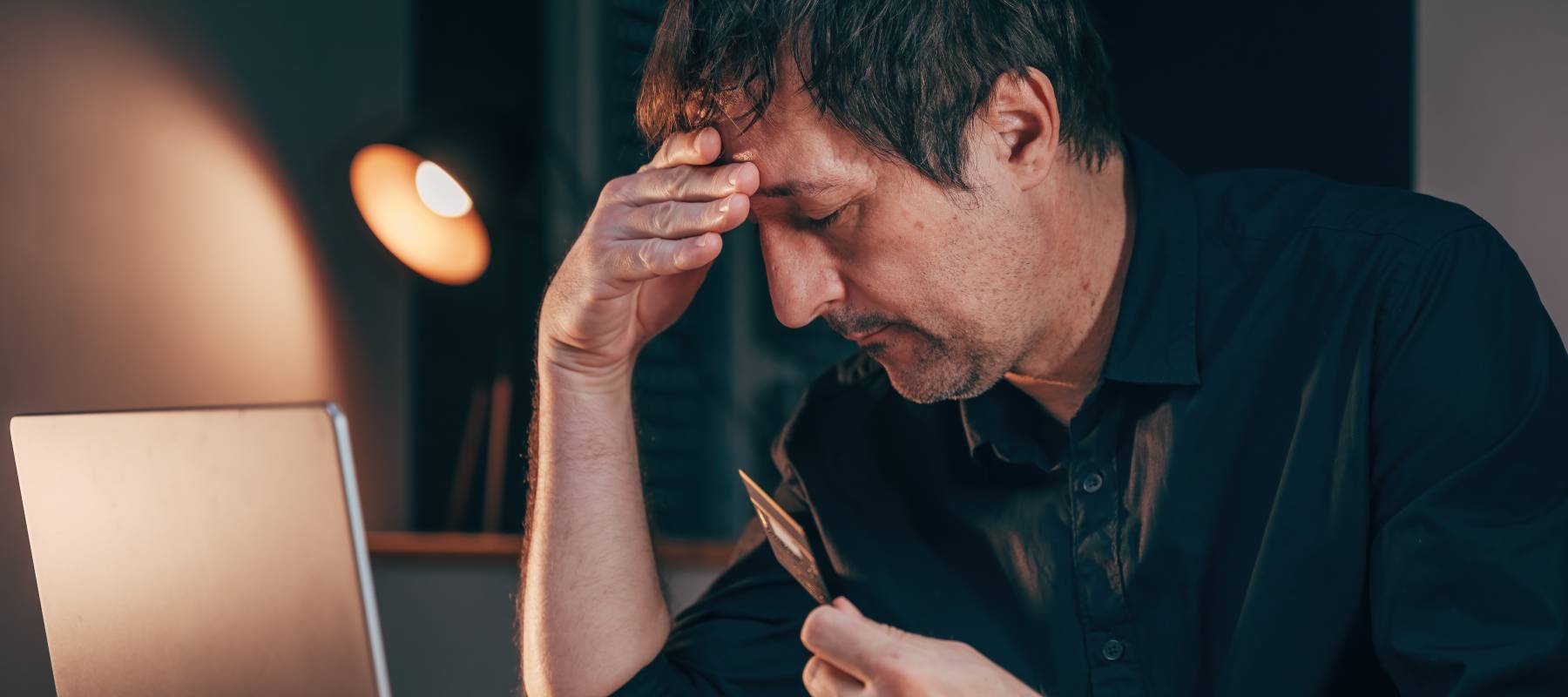 A stressed-looking man grabs his credit card in front of a laptop.