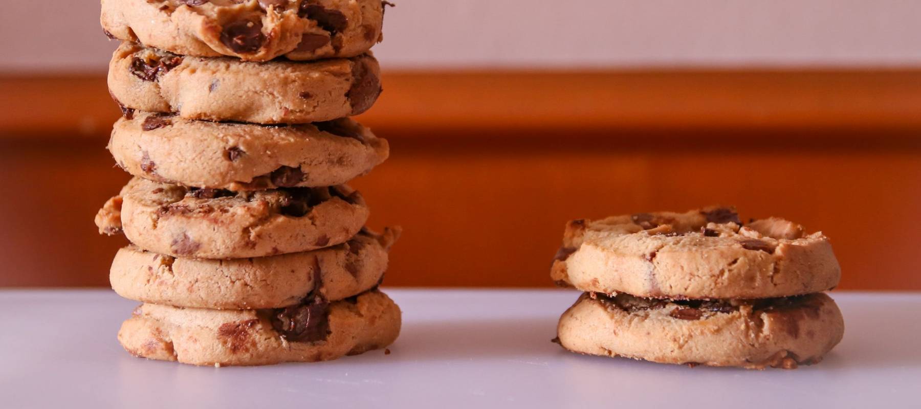 Close up wallpaper of two stacks of chocolate chip cookies of unequal height on a flat surface against a brown textured background.