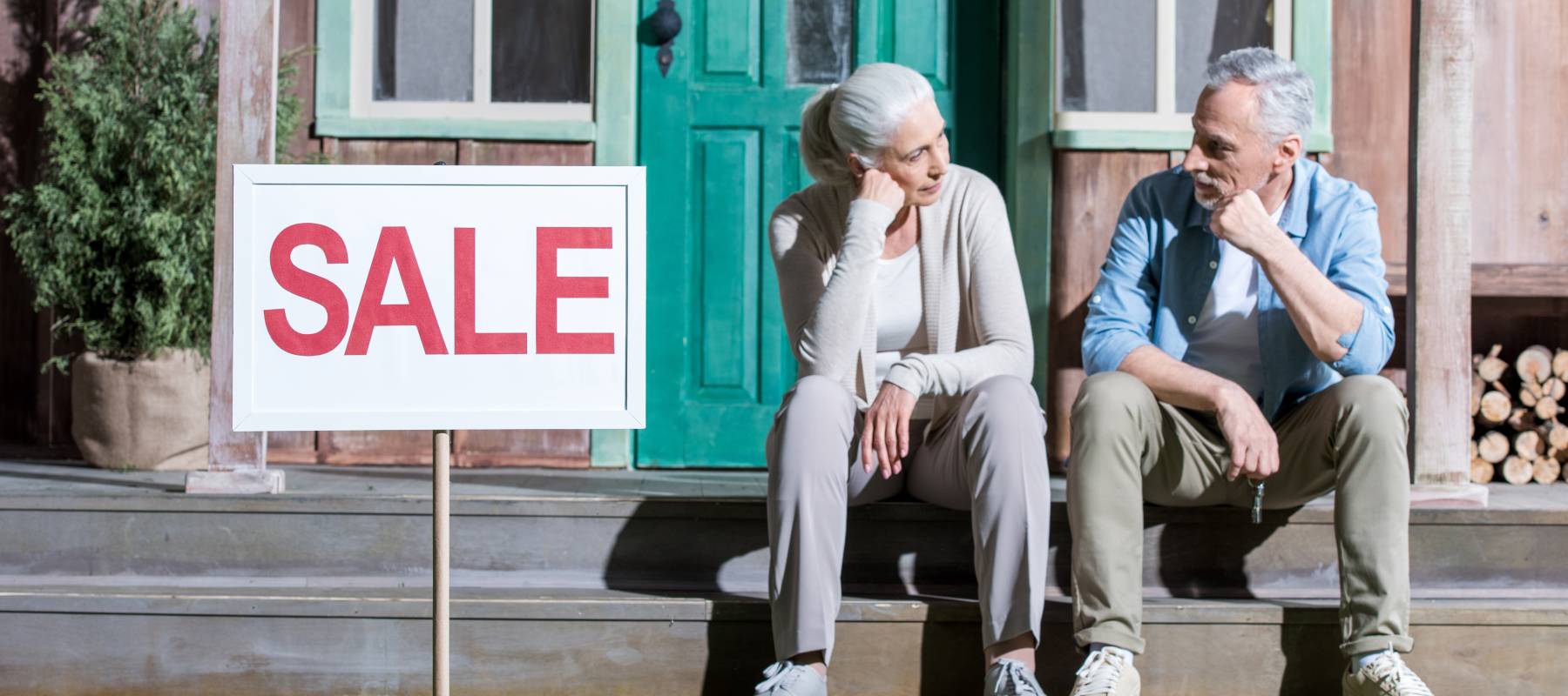 A senior couple sit in front of their home for sale.