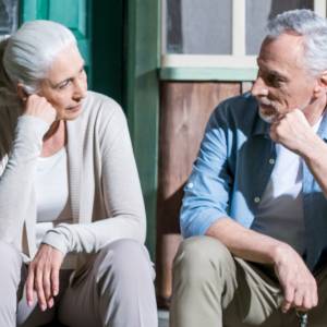 A senior couple sit in front of their home for sale.