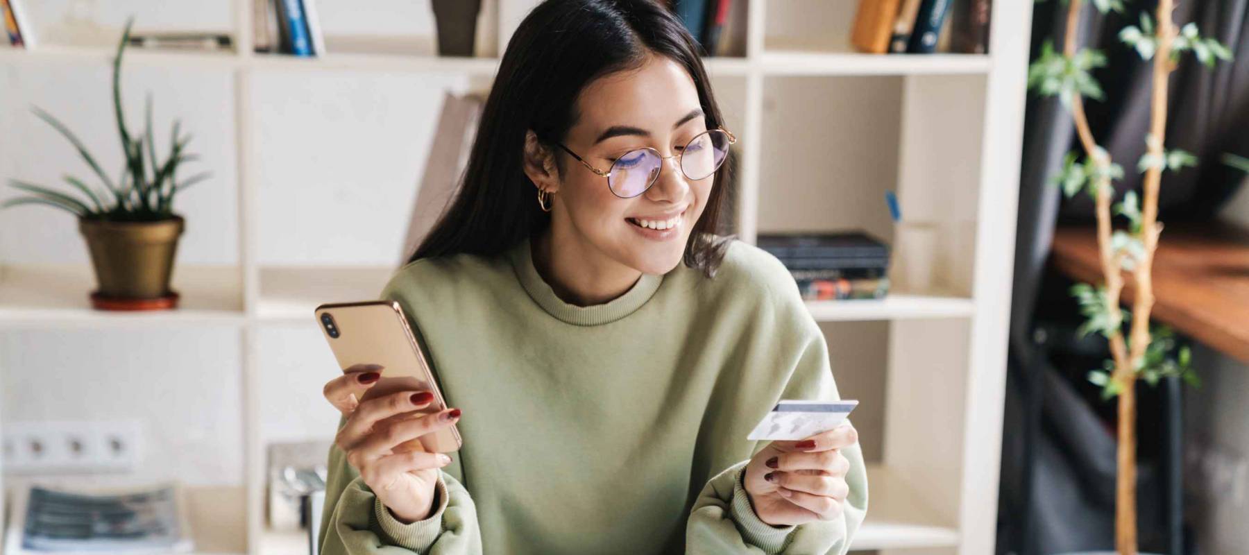 Image of a beautiful happy optimistic young girl student indoors studying using mobile phone holding credit card.