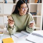 Image of a beautiful happy optimistic young girl student indoors studying using mobile phone holding credit card.