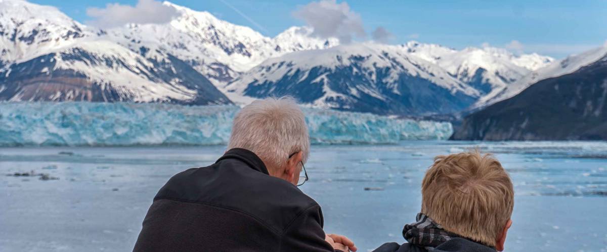 Old couple on a cruise ship in Alaska. Husband and wife are enjoying an amazing view of Hubbard Glacier and iceberg. Snow peaks and ice all around in the sea.