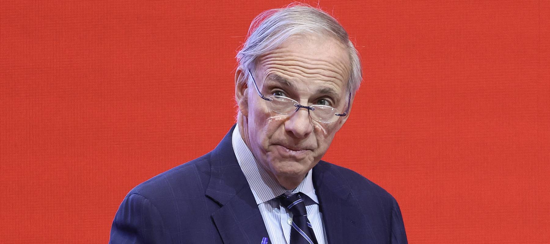 Ray Dalio speaks into a microphone and gestures with one hand against a bright red backdrop.
