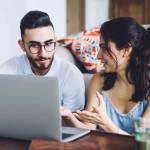Intelligent couple sitting together on floor at table with laptop while woman showing freelance work results on laptop screen and gesticulating with open palms on background with sofa