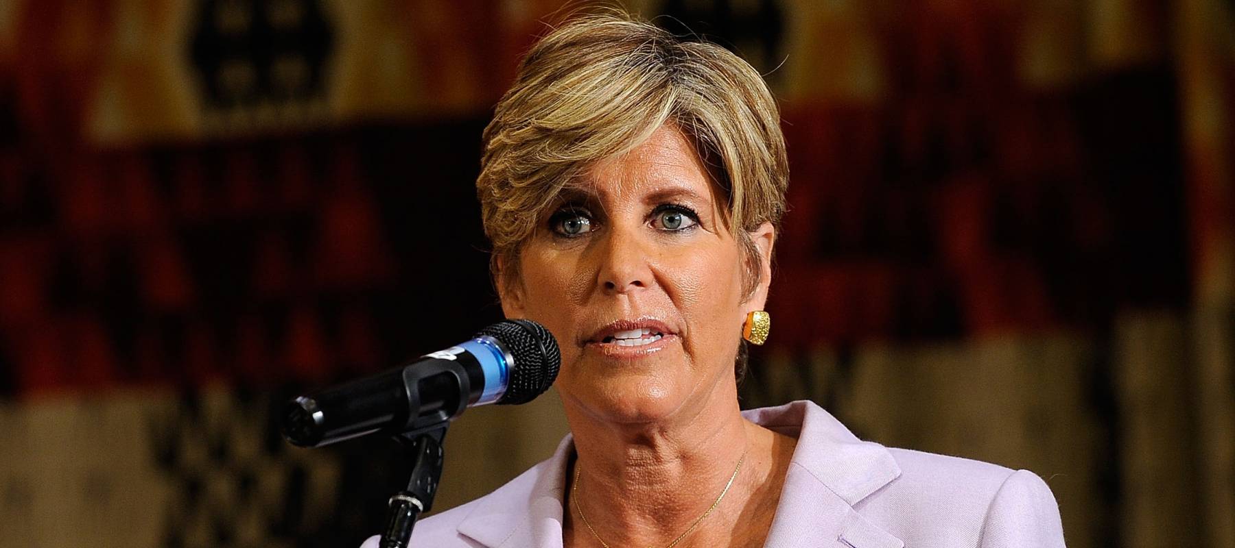 Finance icon Suze Orman gestures broadly while wearing a lavender blazer.