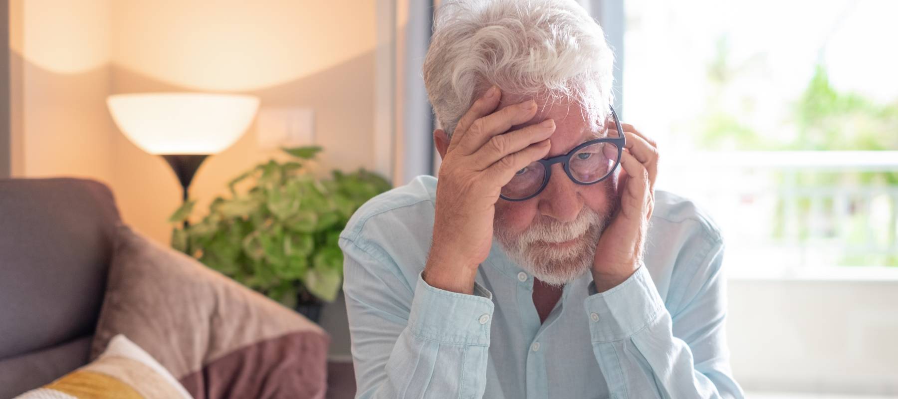 Older bespectacled man sitting alone at home staring into space, old man suffering from health problems.