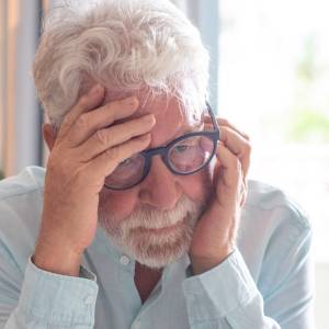 Older bespectacled man sitting alone at home staring into space, old man suffering from health problems.