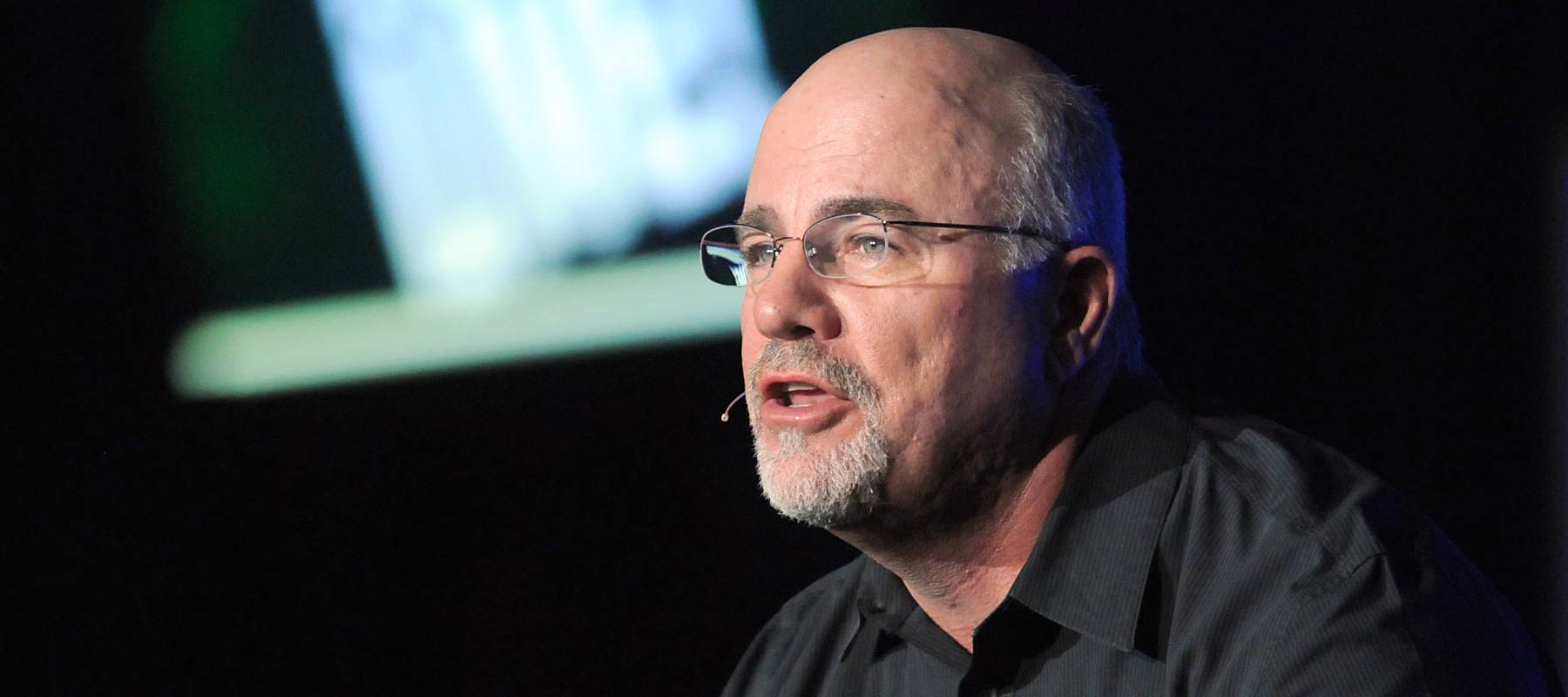 Dave Ramsey, in a black dress shirt, spreads his hands and speaks on stage.