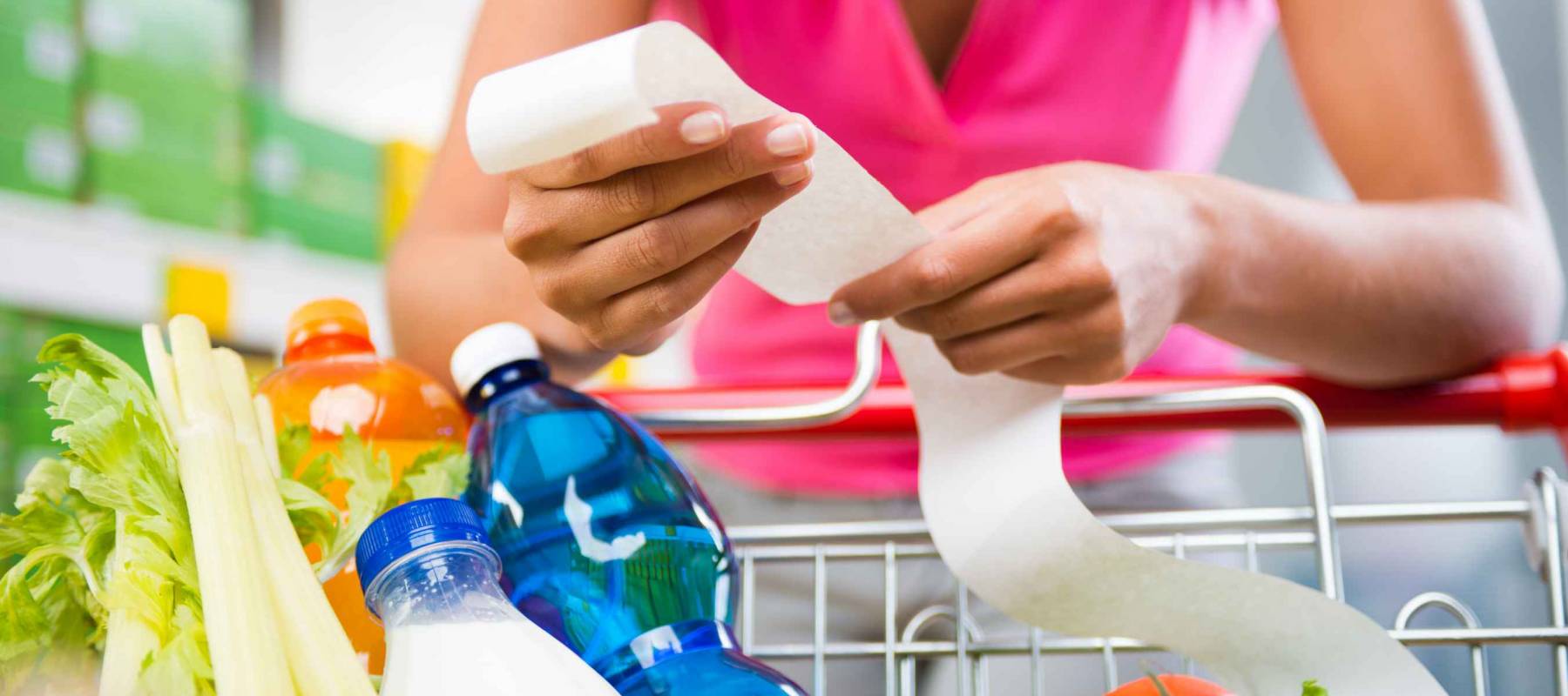 Unrecognizable woman checking a long grocery receipt leaning to a full shopping cart at store.