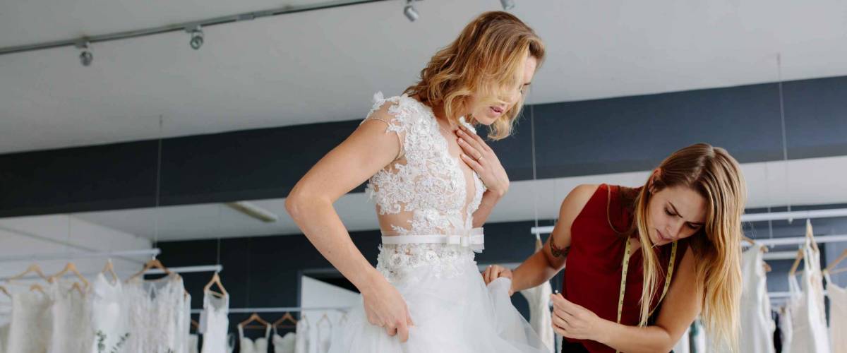 Female making adjustment to wedding gown in fashion designer studio. Bride wearing her wedding gown with female dress designer making final adjustments on dress.