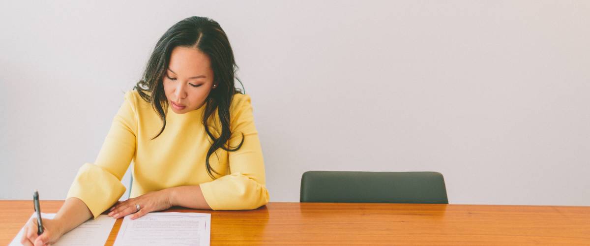 A young, professional asian woman signing papers while sitting at a table in a modern office.