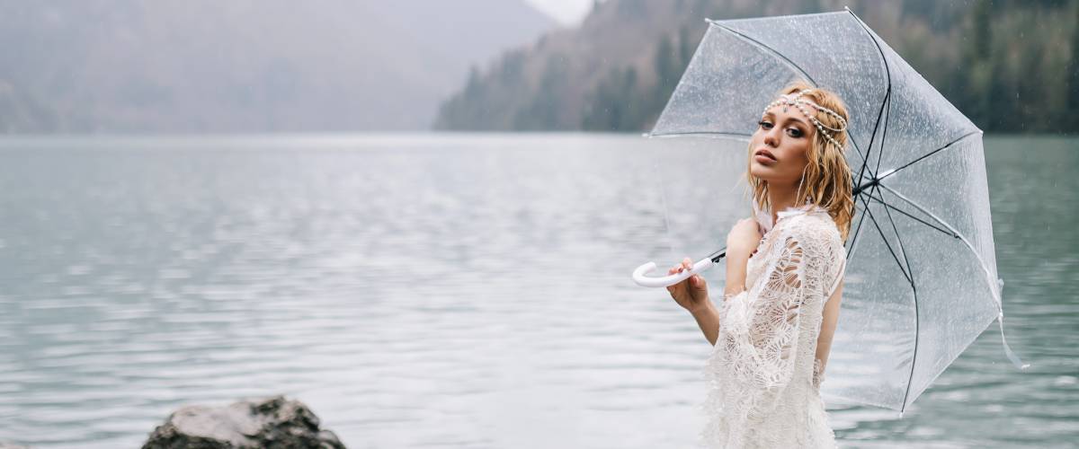 An attractive woman bride in a fashionable wedding dress with an umbrella stands in the rain on sea