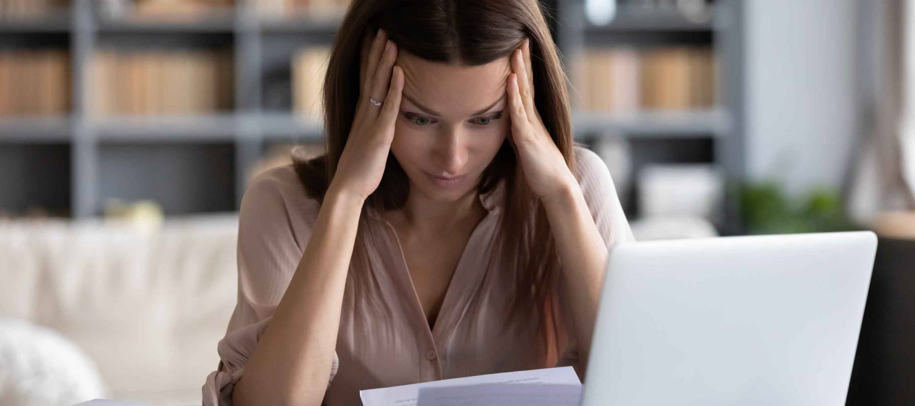 Stressed young woman holding head in hands, looking down at laptop and papers