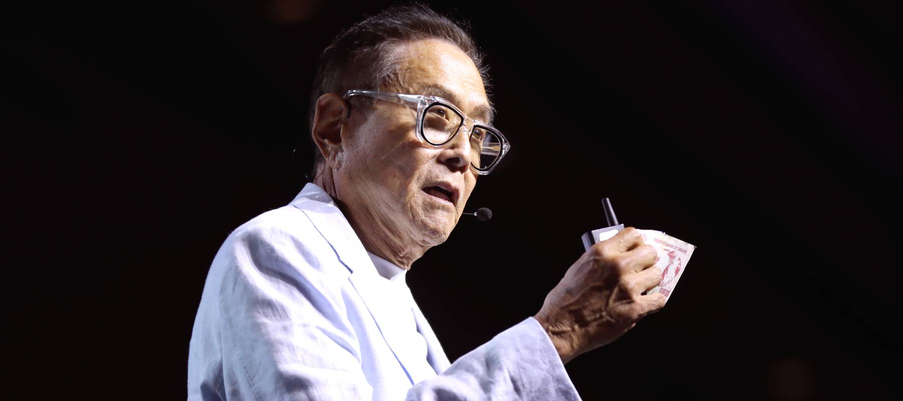 Robert Kiyosaki holds up some bills while wearing a pure white suit