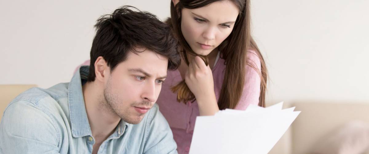 Young serious man holding papers, reading them attentively, sitting with laptop indoors. Anxious woman standing next to him looking at documents. Unpaid domestic bills, checking documentation
