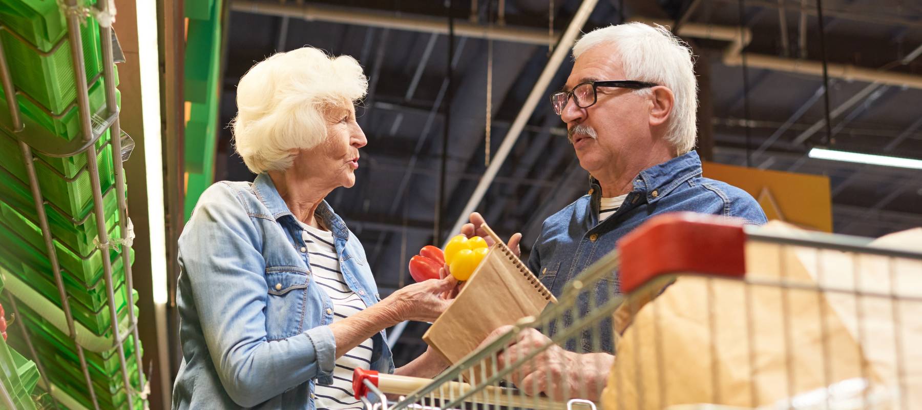 An older couple discuss whether to buy peppers or not in a brightly lit supermarket.