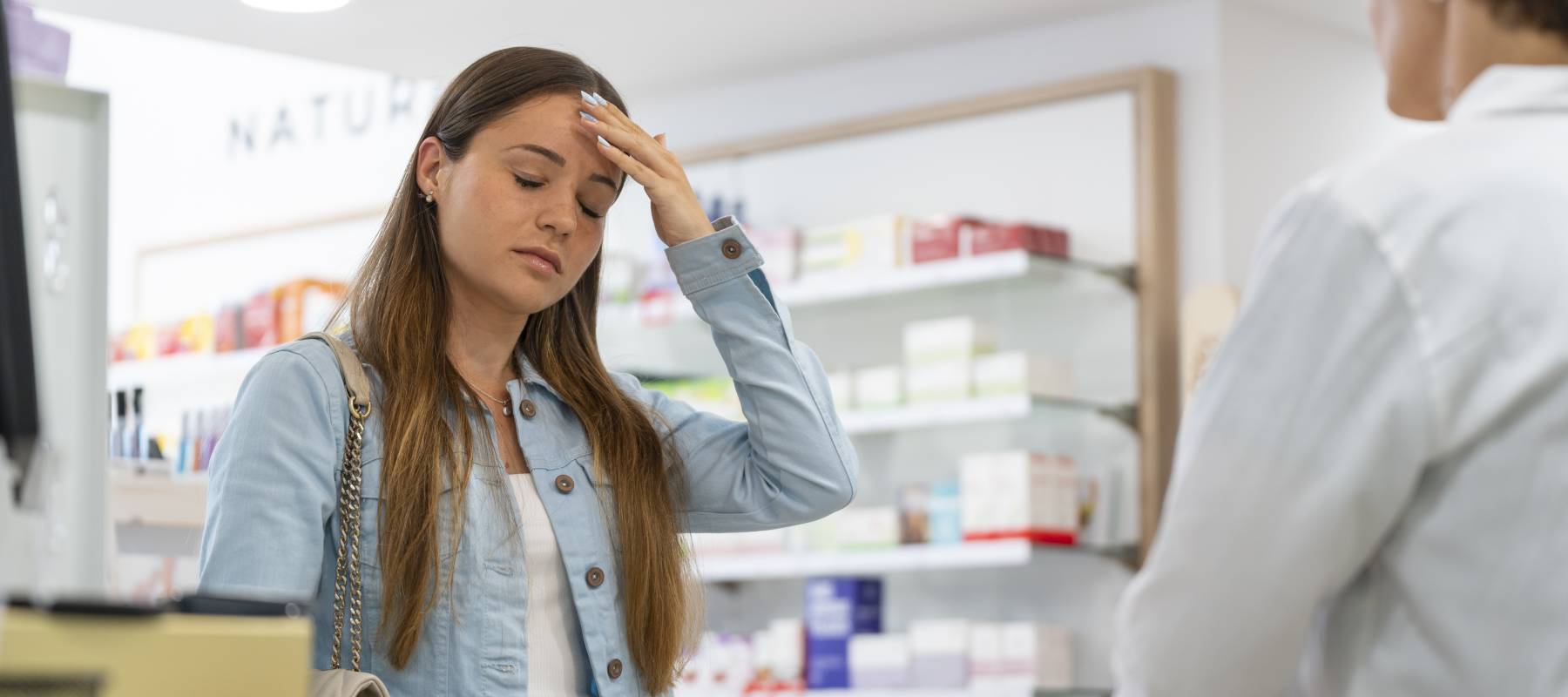 A young woman holds her head in pain at a pharmacy counter.