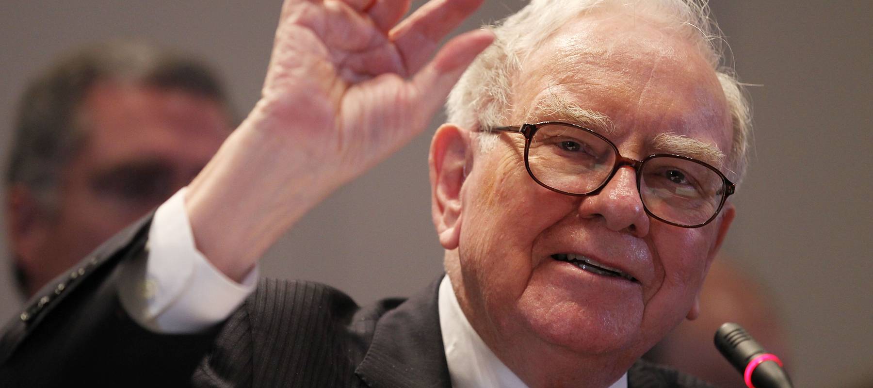 Warren Buffett speaks into a microphone and holds up one hand with his index and thumb slightly apart.