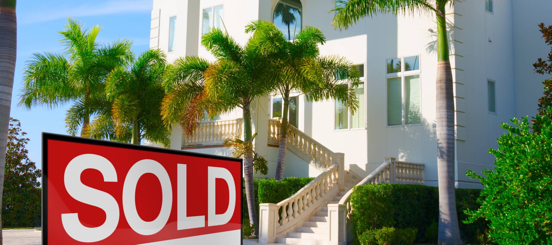 Beautiful purchased luxury residential home with front stairways and lush green palm trees against a blue morning sky with a bright SOLD sign in the front yard.