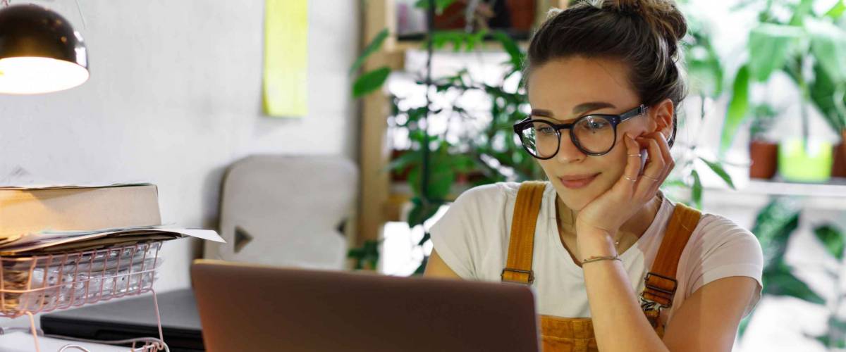 Young woman wearing glasses, works at laptop with hand on her chin