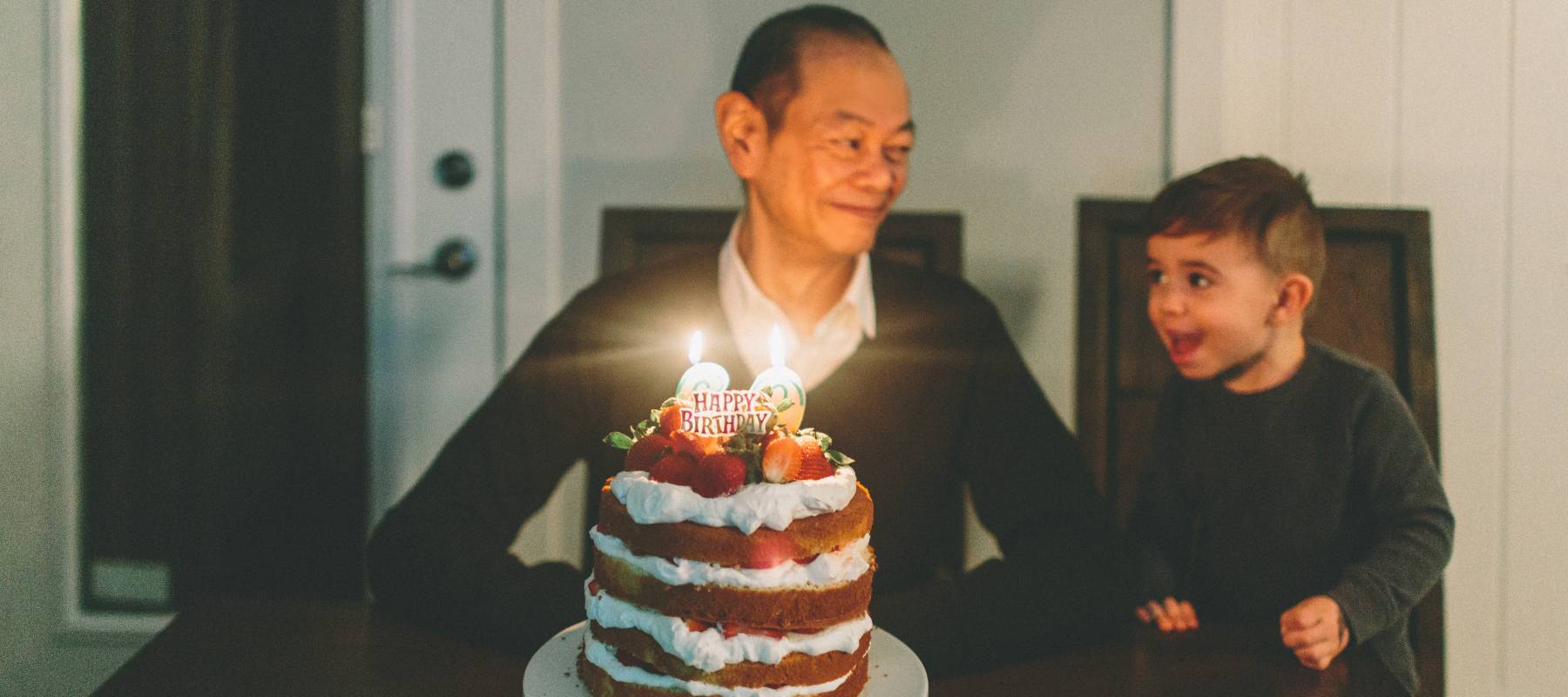 Little boy and older man sit at a table with a big birthday cake in front of them, smiling and looking at each other