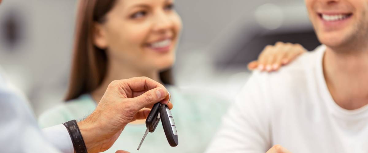 Young couple is smiling while buying a car, middle aged worker of a motor show is giving keys, close-up