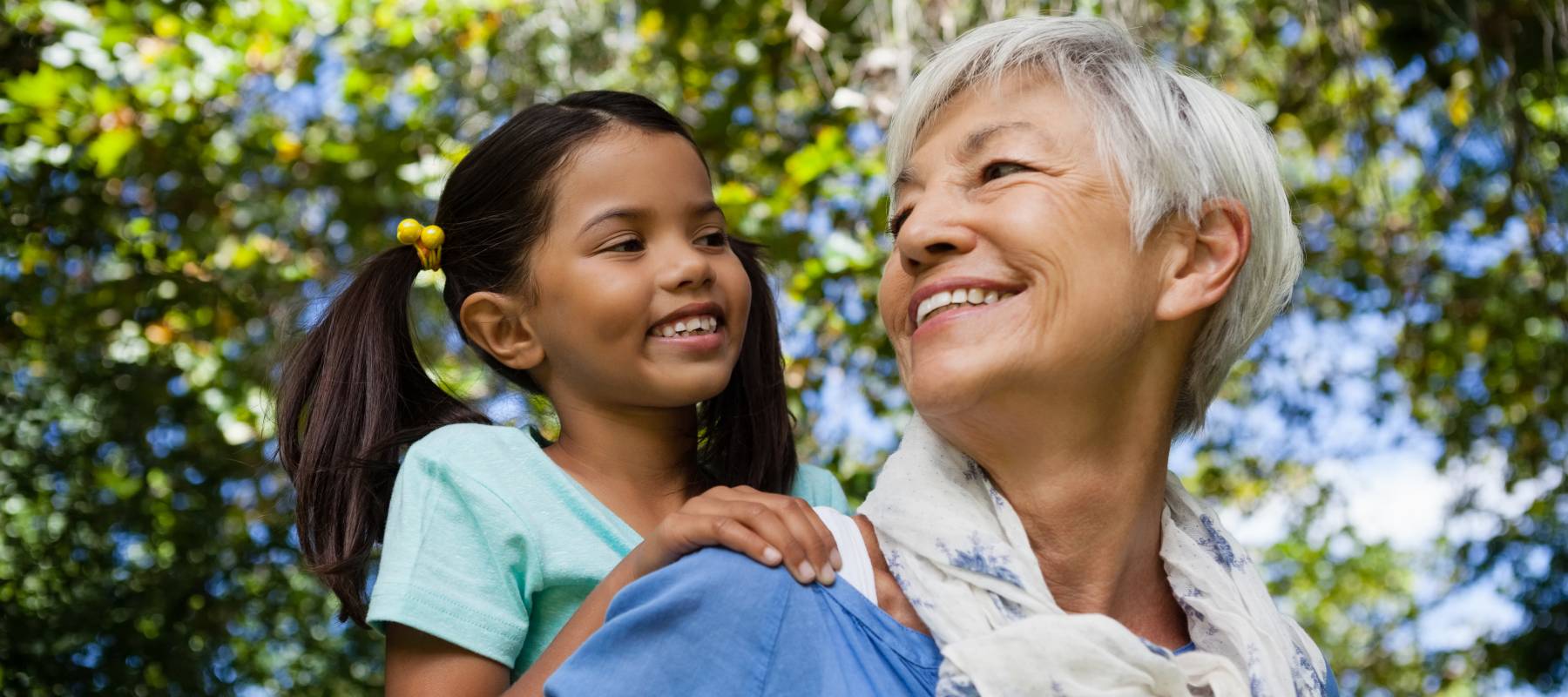 Low angle view of happy grandmother giving piggyback to granddaughter against trees.