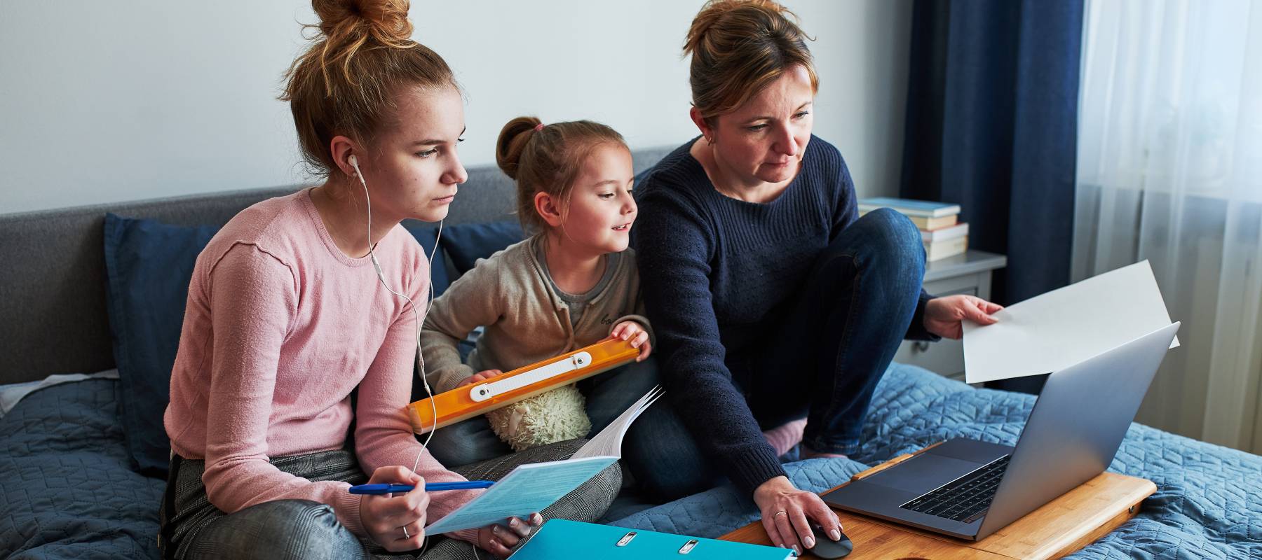 Mother sitting on bed with her two young daughters, looking at laptop together.