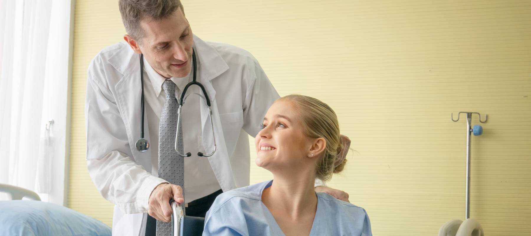 Doctor leaning over in office to talk to young woman in wheelchair, she's smiling up at him