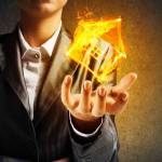 Closeup of businessman in black suit keeping flaming house symbol in hands with dark wall on background.
