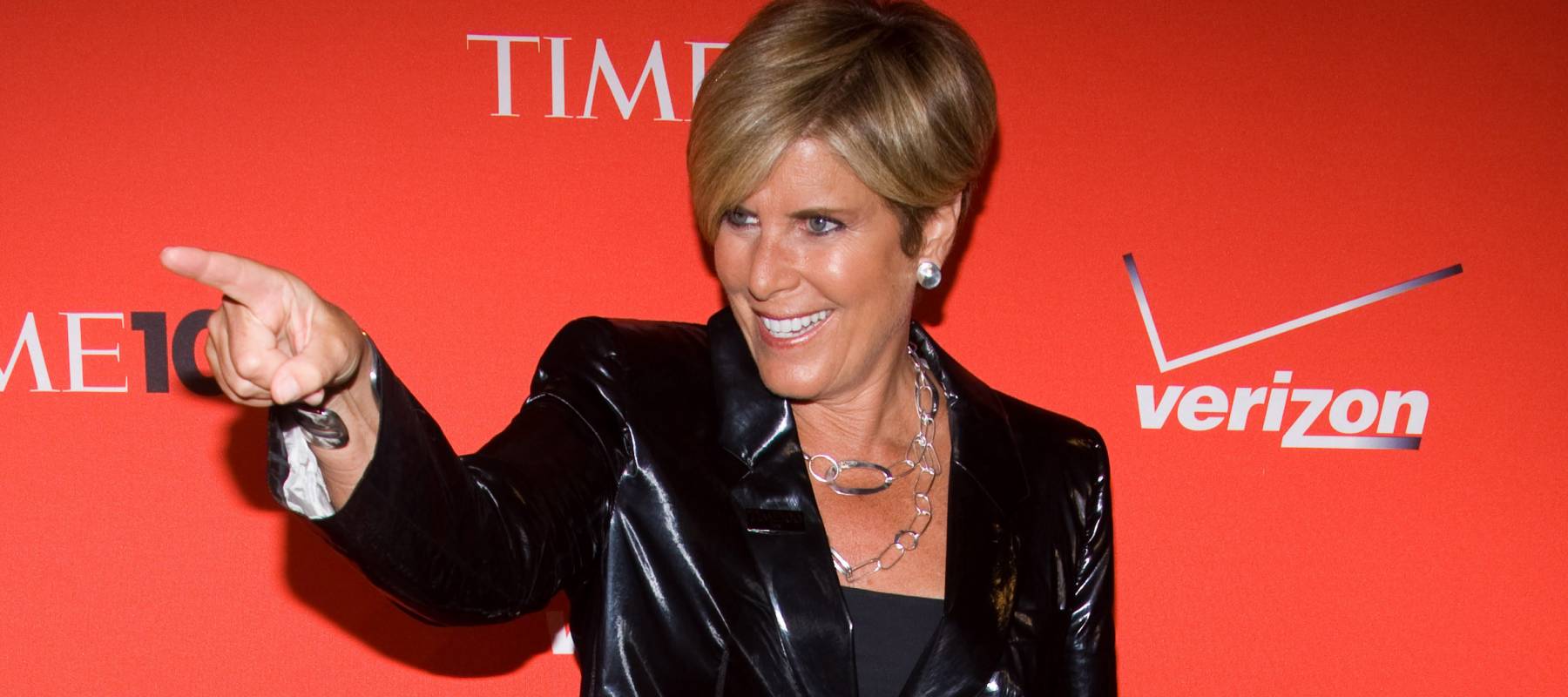 Suze Orman stands in front of a red backdrop with her finger pointing