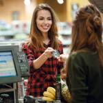 Image of smiling young lady standing in supermarket shop near cashier's desk holding credit card. Looking aside.