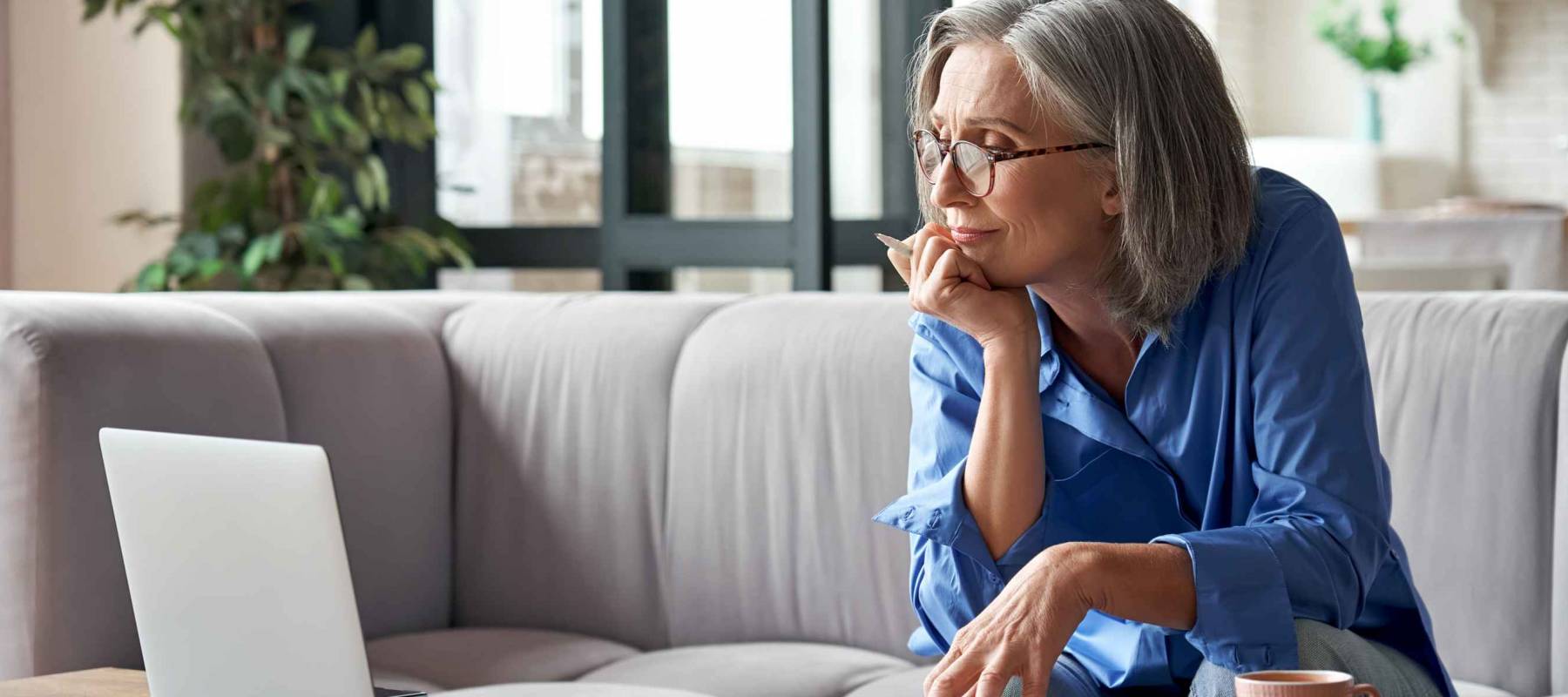 Older woman sits on couch, looking at computer and holding her chin in her hands.