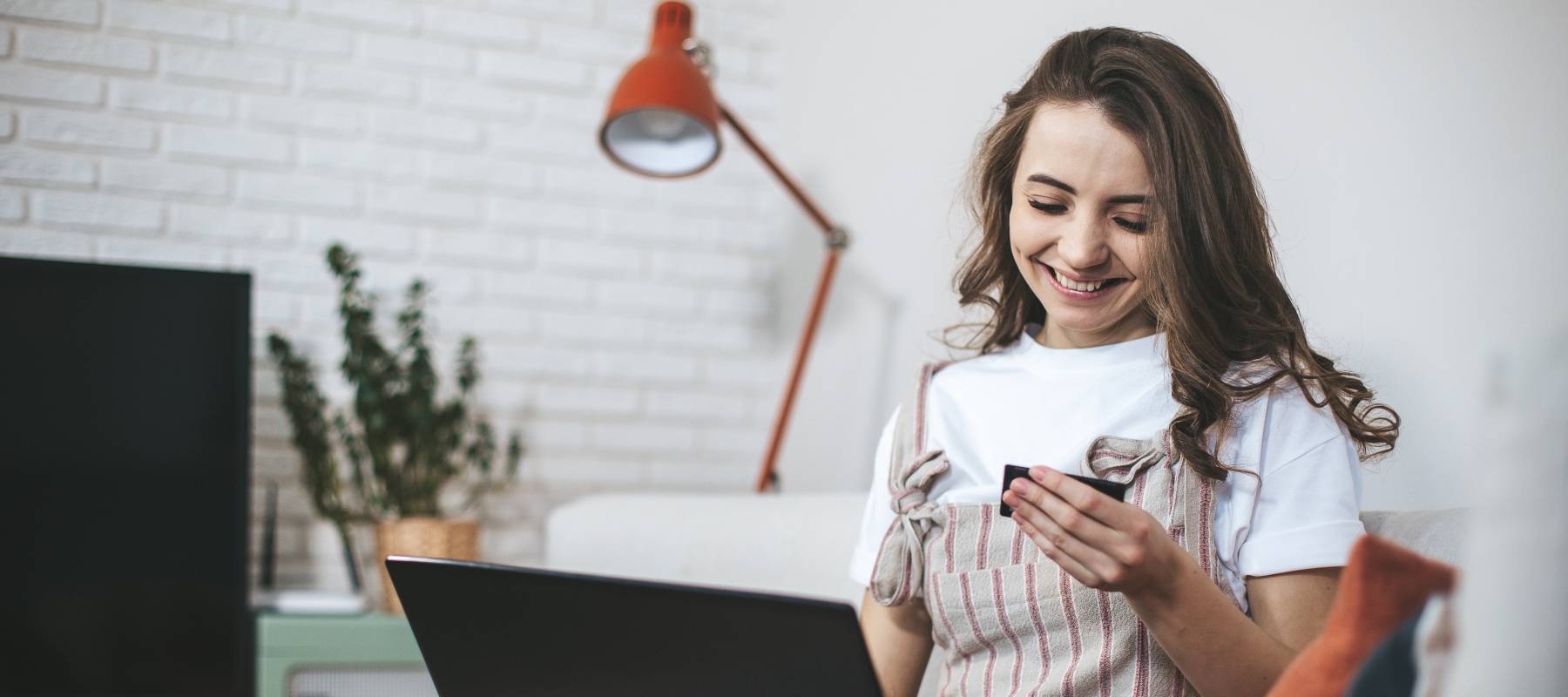 Woman smiles and looks down at a credit card in her hand, sitting at a desk with a laptop in her lap.