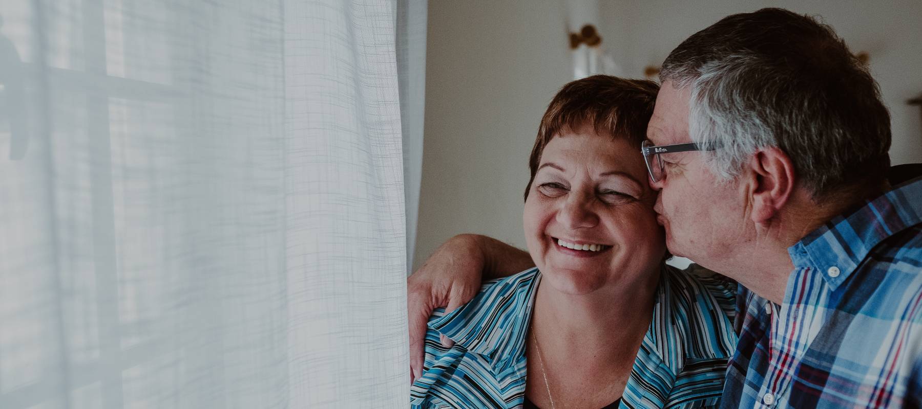 An older couple smiles, posing in front of a window while the man kisses his wife's cheek.