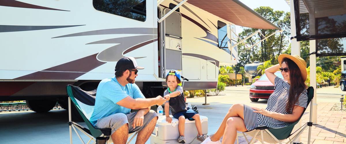 Mother, father and son sitting near camping trailer,smiling.Woman, men, kid relaxing on chairs near car and palms.Family spending time together on vacation near sea or ocean in modern rv park