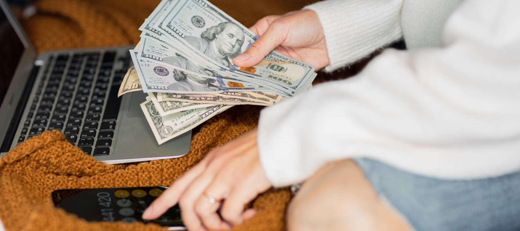 Close up of woman sitting on bed with hand full of cash, using laptop and calculator.