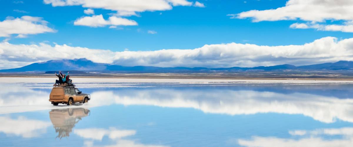 Car with happy people driving on the mirror surface of Salar de Uyuni in Bolivia with clouds reflection