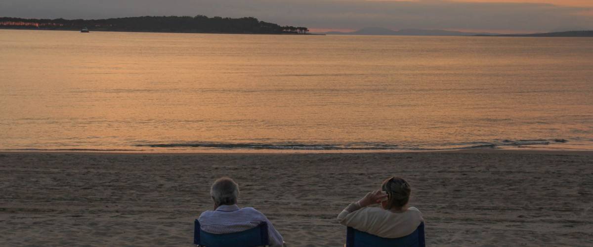 An older couple sit in portable fold-up chairs on the beach to watch the sunset, one talking on mobile phone, in Punta del Este, Uruguay