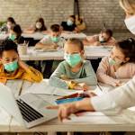 Group of school kids with protective face masks learning on laptop with their teacher during a class in the classroom.