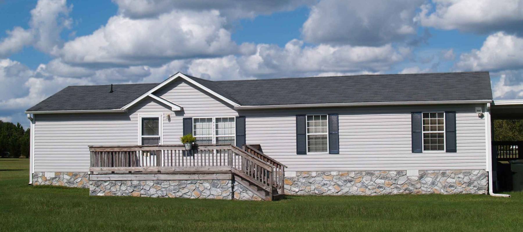 Gray trailer home with stone foundation or skirting and shutters in front of a beautiful sky.