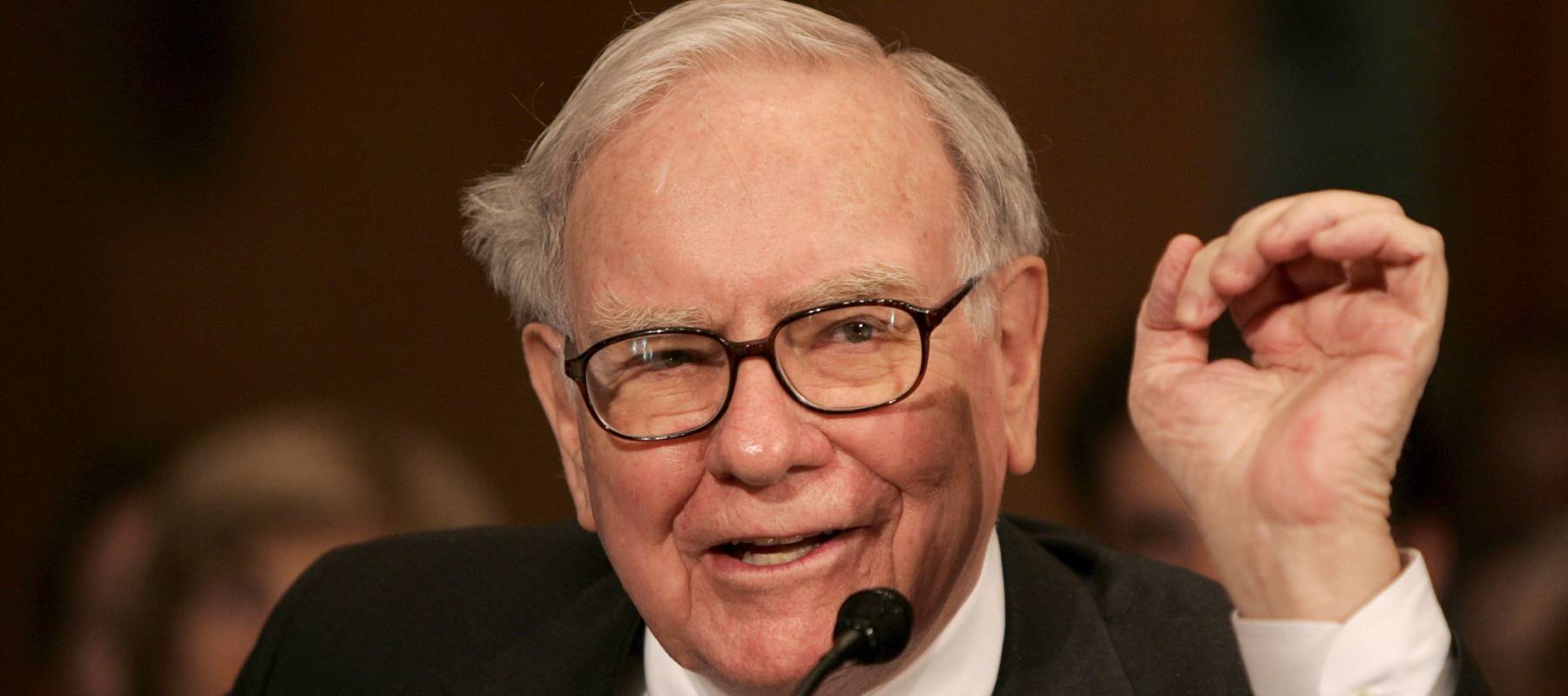Warren Buffett speaks into a microphone and holds his hand up while talking.