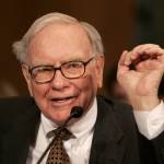 Warren Buffett speaks into a microphone and holds his hand up while talking.